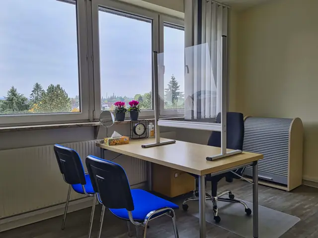 Therapieraum mit Arbeitstisch, blauen Stühlen und Acrylwand vor einer Fensterfront